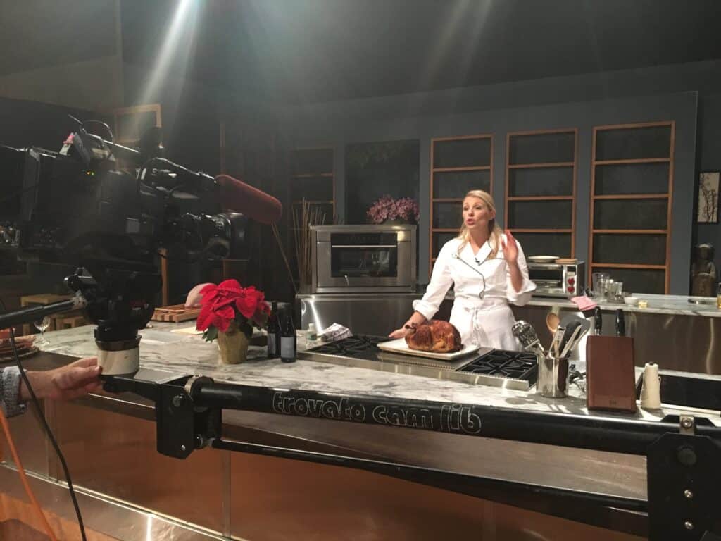 A chef in a white coat presents a cooked roast on a kitchen showroom set featuring Wolf appliances while being filmed by a camera crew.