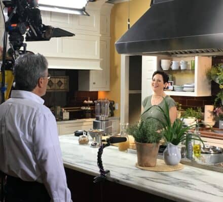 A woman stands in a kitchen showroom set while being filmed by a camera crew, with plants and kitchen items on the counter.