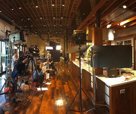 A film crew with cameras and lighting equipment sets up inside a modern kitchen with wooden floors, exposed ceiling beams, and a gleaming Wolf appliance that adds to the luxury kitchen ambiance.