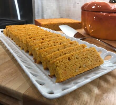Sliced cornbread is neatly arranged on a white rectangular plate; a serving utensil and a pumpkin-shaped dish sit in the background, evoking the warmth of a luxury kitchen appliance showroom.