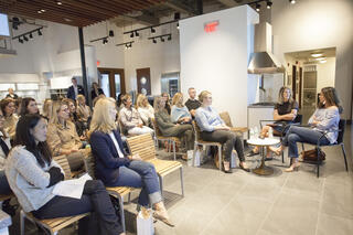 A group of people sits on chairs facing two women having a discussion in a modern, well-lit kitchen showroom with stylish displays.