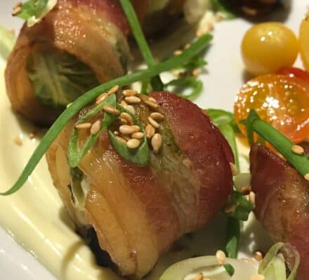 Bacon-wrapped appetizers garnished with green onions and sesame seeds, served on a swirl of creamy sauce with cherry tomatoes—perfect inspiration for your next gathering after browsing a kitchen showroom.