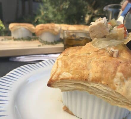 A close-up of a chicken pot pie with golden, flaky crust in a white ramekin on a plate; more pot pies and a drink are in the background, set in an elegant appliance showroom.