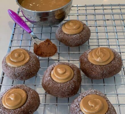 Six chocolate thumbprint cookies topped with caramel sit on a cooling rack in an appliance showroom, with a bowl of caramel sauce and a spoonful of cocoa powder nearby.
