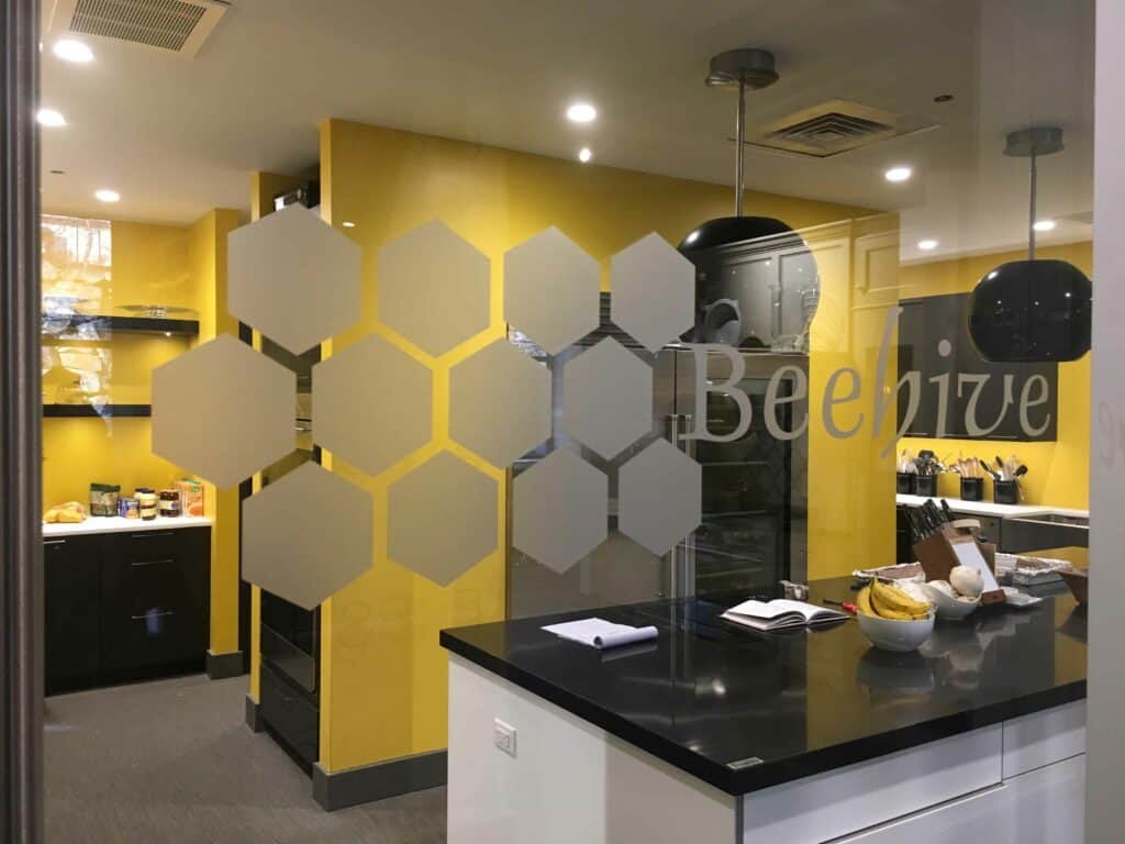 Modern office kitchen with yellow walls, black countertops, and "Beehive" text with hexagonal designs on a glass partition; reminiscent of a stylish kitchen showroom, various kitchen items are visible.