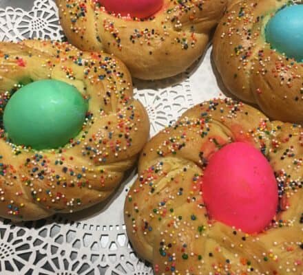 Four braided sweet breads with colorful dyed eggs in the center and multicolored sprinkles on top, beautifully arranged on a doily-lined tray—perfect for showcasing in any luxury kitchen appliance setting or appliance showroom.