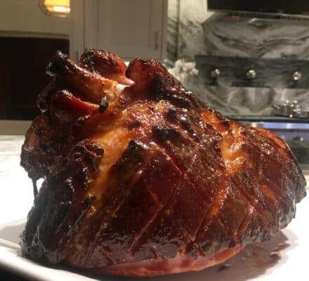A glazed, baked ham with a diamond-cut pattern sits on a white plate in a modern kitchen, showcasing the elegance of a luxury kitchen appliance showroom.