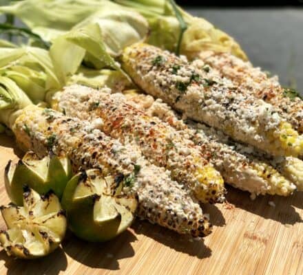 Grilled corn on the cob topped with cheese and spices, prepared using a Wolf appliance, and served on a wooden board with charred lime wedges.