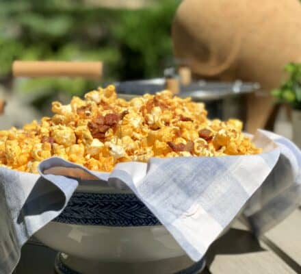 A bowl lined with a cloth is filled with caramel popcorn, sitting on an outdoor table with blurred plants and luxury kitchen appliance accents in the background.