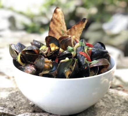A white bowl filled with cooked mussels, garnished with herbs and sliced red peppers, sits beside two pieces of toasted bread—an elegant dish perfect for enjoying in a luxury kitchen appliance showroom.