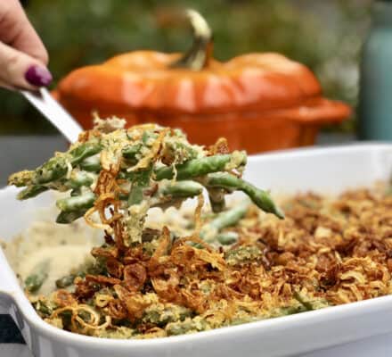 A hand serves green bean casserole topped with crispy fried onions from a white dish in a kitchen showroom; a pumpkin-shaped dish is blurred in the background, highlighting premium Wolf and Sub-Zero appliances.