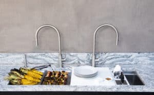A marble countertop with two sinks, plates stacked by one sink, utensils, grilled corn, skewers, and tongs arranged alongside in an elegant appliance showroom.