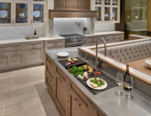Modern kitchen with an island countertop featuring plated food, bread, grapes, wine glasses, and a bottle of wine; luxury kitchen appliances and glass-front cabinets in the background evoke an upscale appliance showroom feel.