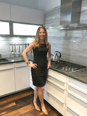 A woman in a black dress stands and smiles in a modern kitchen showroom with white cabinets, a gas stove, and marble backsplash, showcasing the latest sub zero appliance.