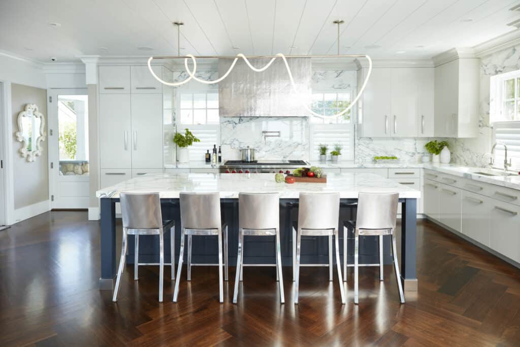 Modern kitchen with white cabinets, marble countertops, luxury kitchen appliances, an island with five metal chairs, and a sculptural light fixture overhead.