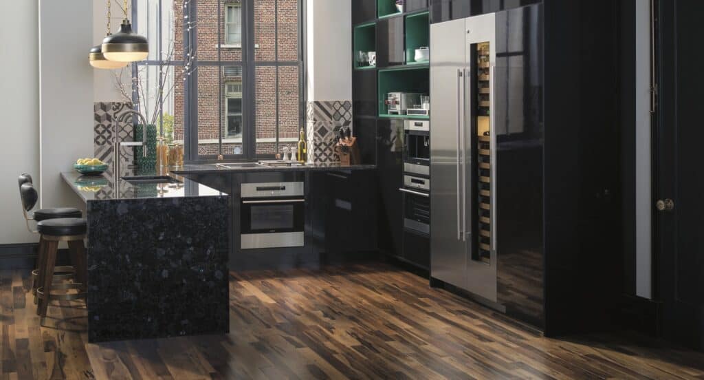 Modern kitchen with black countertops, stainless steel appliances, a large refrigerator, and wooden flooring. Large windows let in natural light—perfect inspiration for any kitchen showroom.