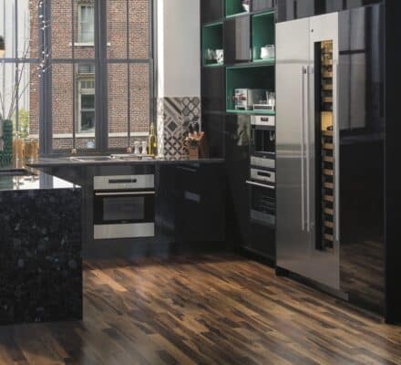 Modern kitchen with black countertops, stainless steel appliances, a large refrigerator, and wooden flooring. Large windows let in natural light—perfect inspiration for any kitchen showroom.