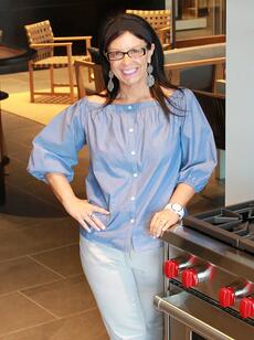 A woman wearing glasses, a blue off-shoulder blouse, and white pants stands and smiles next to a Wolf appliance with red knobs in a modern kitchen.
