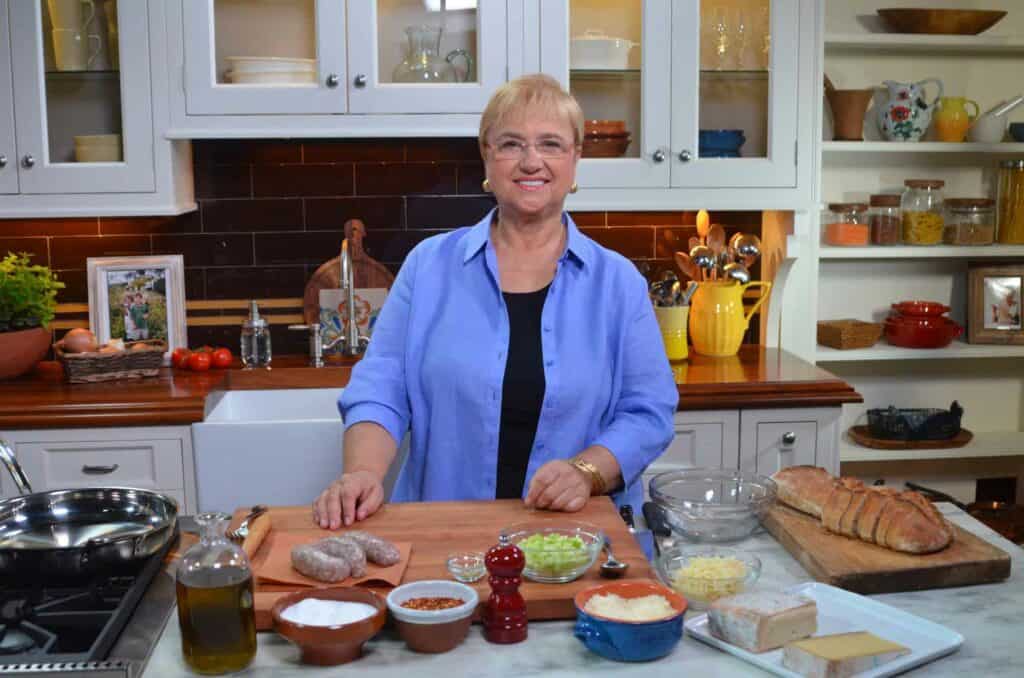 A woman stands in a kitchen showroom behind a counter with bread, seasonings, cheese, oil, and other ingredients. Shelves with dishes and jars are in the background, and a luxury Sub Zero appliance adds elegance to the space.