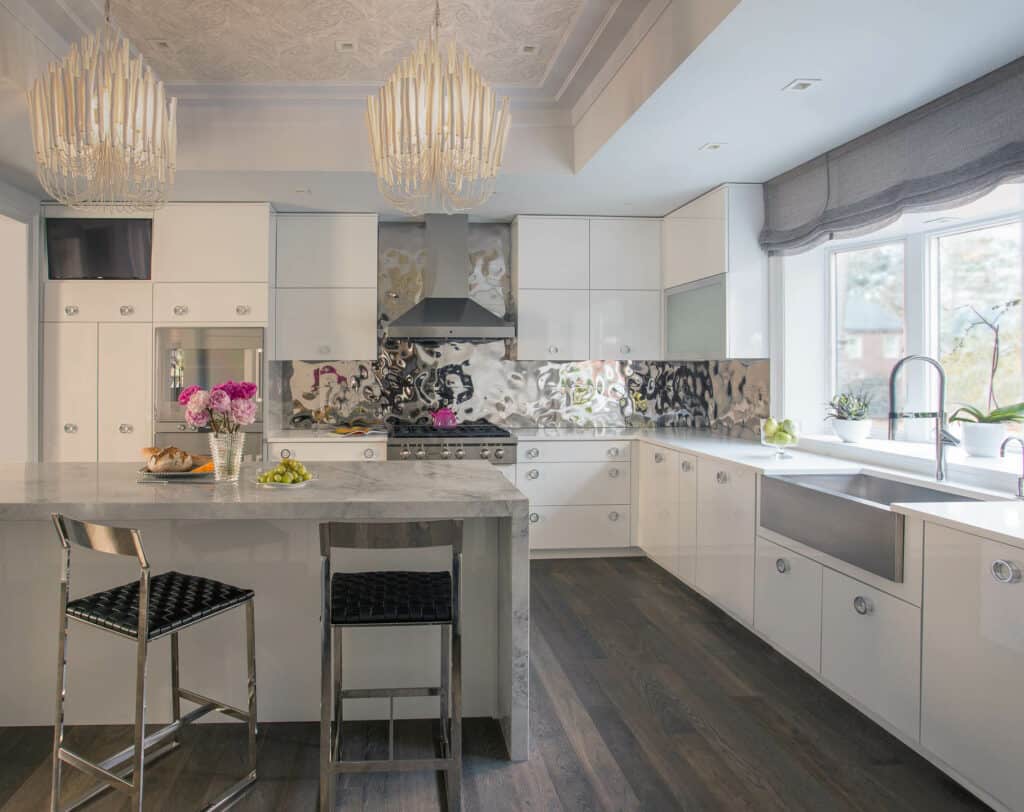 Modern kitchen with white cabinets, a marble island, two metal bar stools, stainless steel and Sub Zero appliances, large sink, and two pendant lights. Flowers and fruit add color to this luxury kitchen appliance showcase.