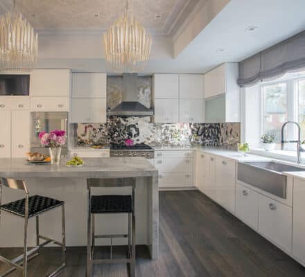 Modern kitchen with white cabinets, a marble island, two metal bar stools, stainless steel and Sub Zero appliances, large sink, and two pendant lights. Flowers and fruit add color to this luxury kitchen appliance showcase.
