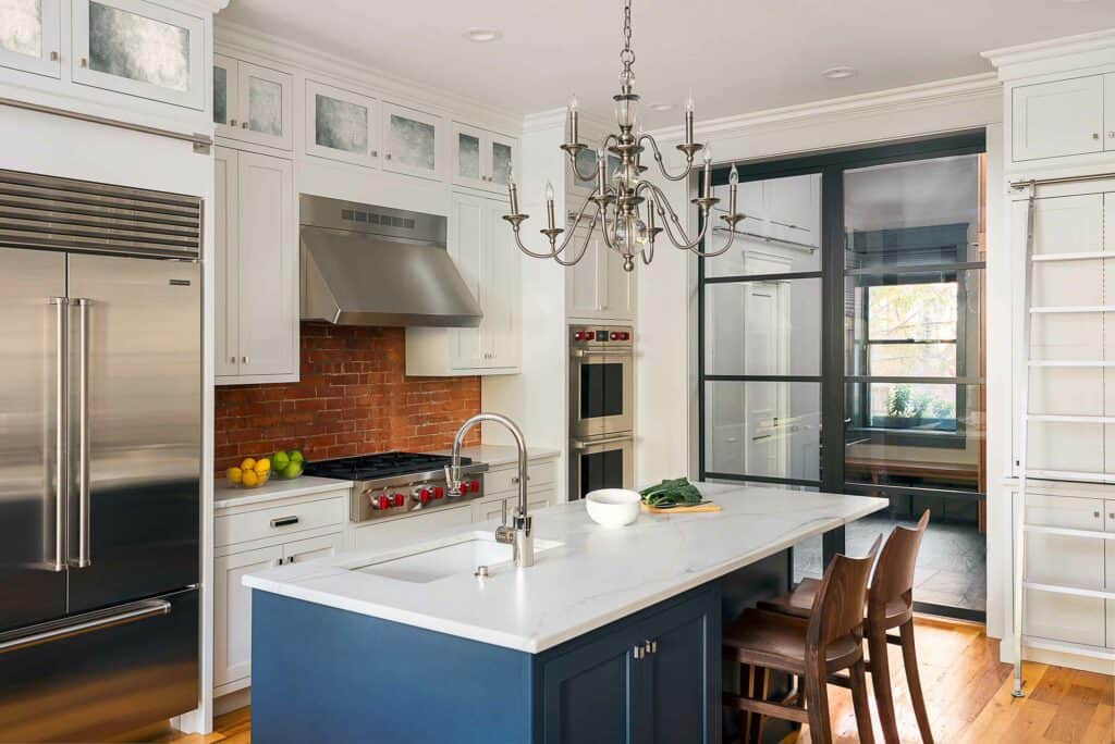 Modern kitchen with white cabinets, stainless steel and Wolf appliances, a brick backsplash, blue island with marble countertop, and a chandelier above the island with two wooden chairs.