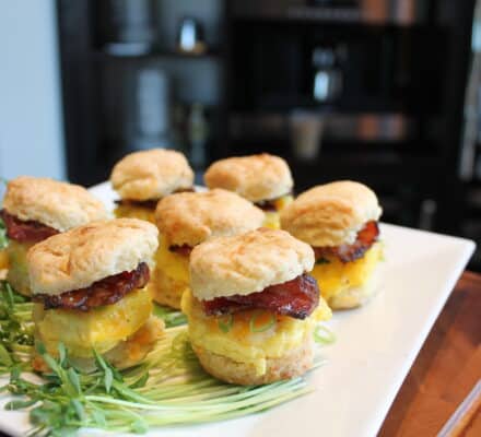 A white plate with mini biscuit sandwiches filled with egg, bacon, and cheese, garnished with fresh sprouts, set on a wooden surface in an elegant appliance showroom.
