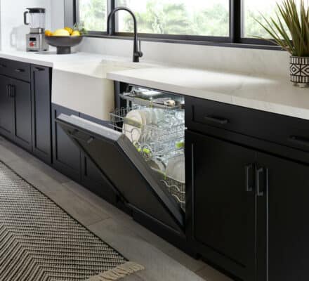 A modern kitchen with black cabinets, a white countertop, and a luxury kitchen appliance dishwasher partially open, showing clean dishes inside. A rug lies on the floor, and a plant and fruit bowl sit on the counter.