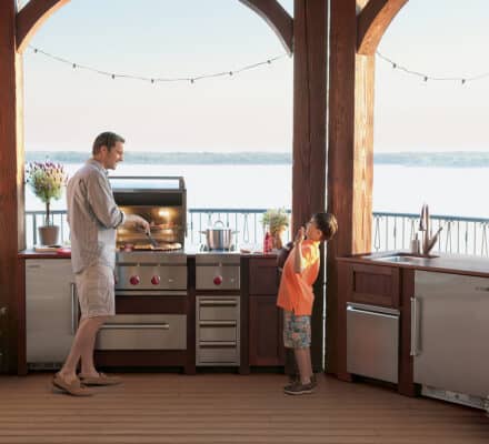 An adult grills food while a child stands nearby on a covered outdoor patio with a lake view and luxury kitchen appliances built into the outdoor space.
