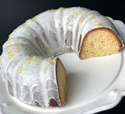 A lemon-glazed bundt cake with a slice removed, displayed on a white decorative plate against a dark background, evokes the elegance of a luxury kitchen appliance collection in a modern kitchen showroom.