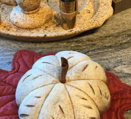 A decorative white pumpkin with brown accents rests on a red leaf-shaped mat atop a granite countertop in a kitchen showroom, with cork coasters and a glass candle holder in the background.