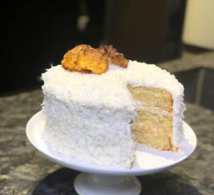 A round, white frosted layer cake with a slice removed, topped with dried pineapple pieces, beautifully displayed on a white cake stand in a modern kitchen showroom.