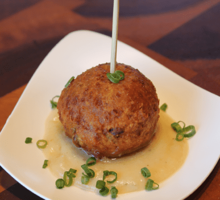 A fried, round croquette sits on a white sauce, garnished with chopped green onions, on a white plate with a wooden skewer—perfectly styled for a kitchen showroom display.