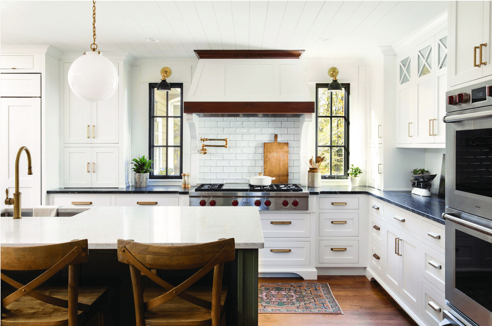 Modern kitchen with white cabinets, black countertops, stainless steel appliances including a Sub Zero appliance, a central island with wooden stools, brass fixtures, and two windows flanking a tiled backsplash.