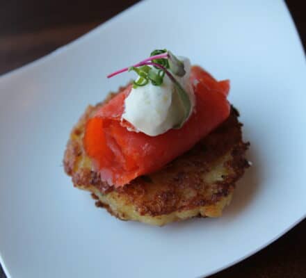 A plate with a potato pancake topped with smoked salmon, a dollop of cream, and microgreens, served on a white dish—an elegant bite you'd expect from an appliance showroom featuring Sub Zero appliances.