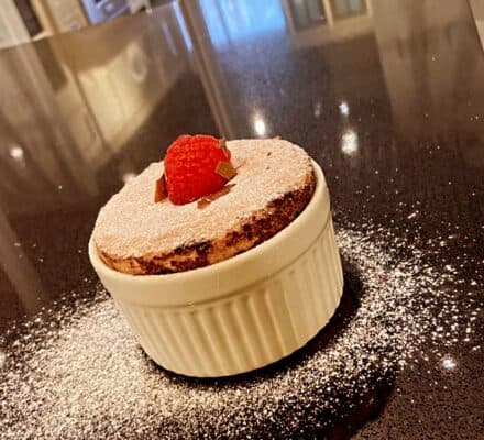 A chocolate soufflé in a white ramekin, topped with a raspberry and dusted with powdered sugar, sits on a dark kitchen countertop—showcasing the artistry possible with a Wolf appliance.