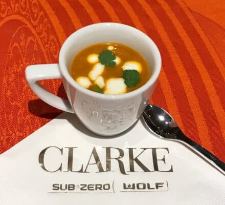 A white cup of orange soup garnished with cream and cilantro sits on a Clarke-branded napkin beside a metal spoon, on an orange tablecloth—evoking the warm elegance of a luxury kitchen appliance display.