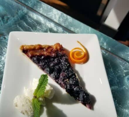 A slice of blueberry tart with whipped cream, a mint leaf, and an orange peel twist on a square white plate—perfect for serving in any luxury kitchen appliance showcase or elegant kitchen showroom.