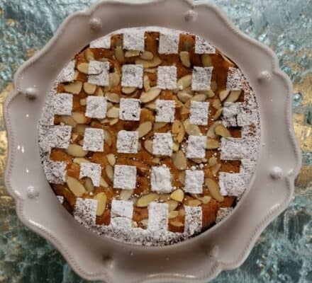 A round cake with a powdered sugar checkerboard pattern and sliced almonds on top, served on a white scalloped plate over a textured glass surface—perfectly baked using a luxury kitchen appliance.