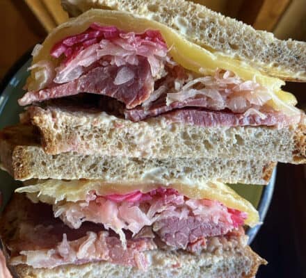 A close-up of a stacked sandwich with corned beef, sauerkraut, cheese, and sauce between slices of rye bread, served fresh in a kitchen showroom featuring premium Sub Zero appliances.