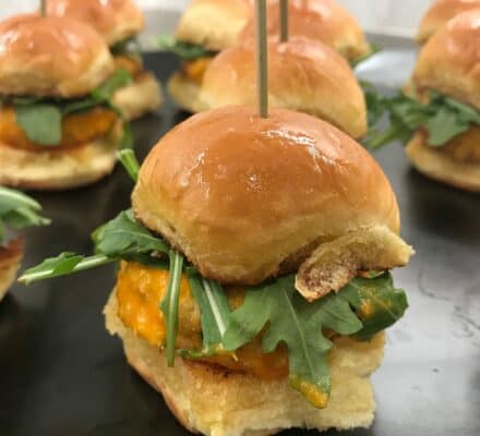 Mini sandwiches with cheese, arugula, and breaded patties on shiny buns, each secured with a toothpick, are artfully arranged on a dark tray—perfect bites for your next kitchen showroom gathering.
