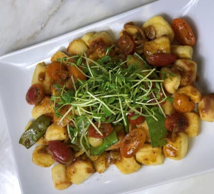 A rectangular plate of gnocchi topped with cherry tomatoes, snap peas, microgreens, and a brown sauce, beautifully presented on a marble surface in an elegant kitchen showroom.