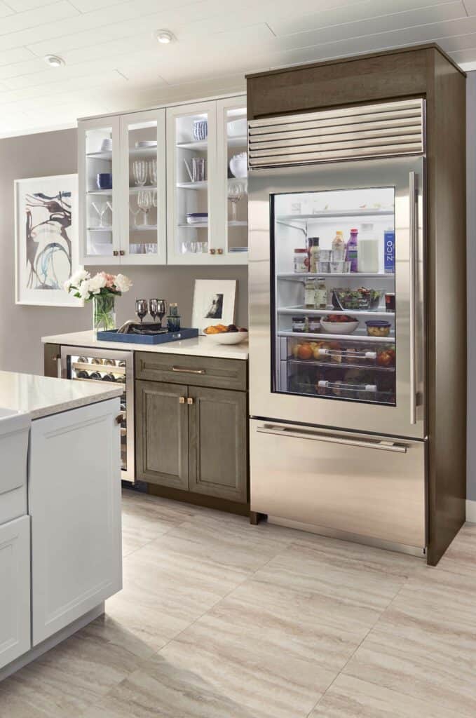 Modern kitchen with a glass-door refrigerator displaying organized food items, adjacent to cabinets, countertop, and a wine cooler. This luxury kitchen appliance setup resembles a high-end kitchen showroom, with art and flowers in the background.