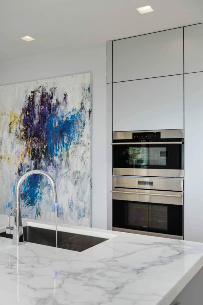 Modern kitchen with a marble countertop, built-in Wolf appliance ovens, a tall faucet, and an abstract painting with blue, purple, and yellow accents on the wall—perfect inspiration for any luxury kitchen appliance showroom.