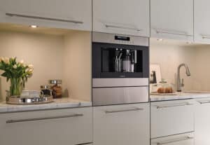 Built-in coffee machine in a modern kitchen, surrounded by white cabinets and marble countertops with flowers, a tray, and a cake stand. This luxury kitchen appliance adds elegance to any space.