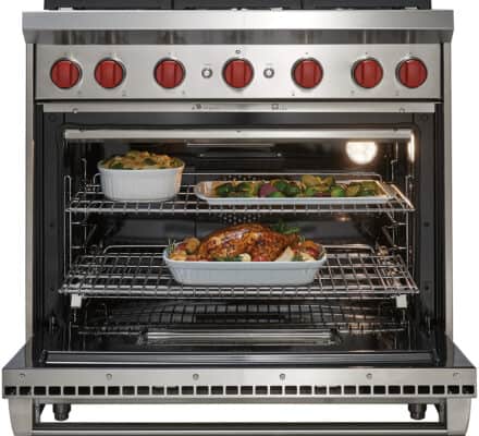 A stainless steel oven with red knobs, a standout luxury kitchen appliance, is open to reveal two trays of cooked food on racks inside, including a roasted chicken and assorted vegetables.