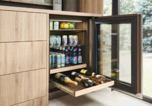 A built-in mini fridge, displayed in an appliance showroom with its glass door open, reveals shelves stocked with cans, bottles, lemons, limes, and a pull-out drawer filled with wine bottles.