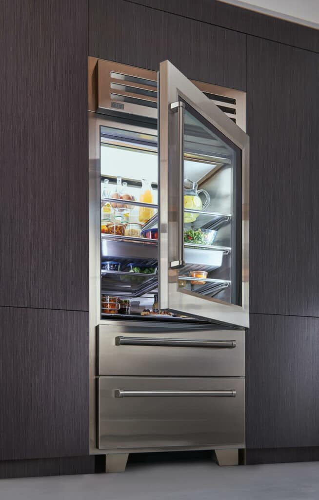 A stainless steel refrigerator with the door open, showing shelves stocked with food and drinks. This luxury kitchen appliance is built into dark wood cabinetry, creating an elegant display fit for an appliance showroom.