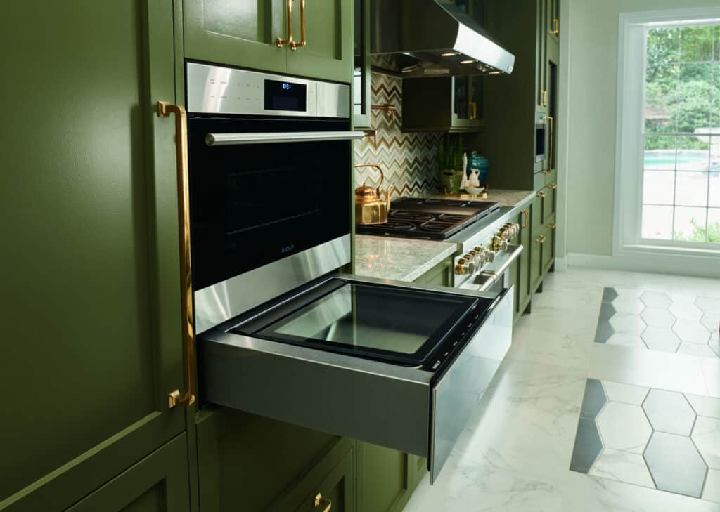 A modern kitchen showroom with green cabinets, a Wolf appliance wall oven open, and a gas cooktop, overlooking a window with a pool view.