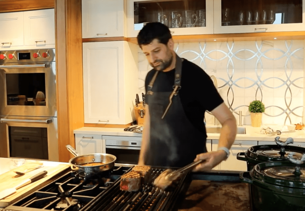 A man wearing an apron is grilling meat on a stovetop grill in a modern kitchen showroom with white cabinets and sleek Sub Zero appliances.